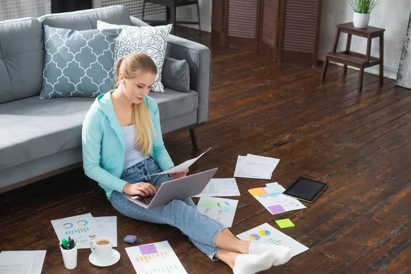 Jovem trabalha com documentos usando um laptop enquanto está sentado no chão em casa. Estudante, empreendedora ou freelancer trabalhando ou estudando remotamente via internet. — Fotografia de Stock