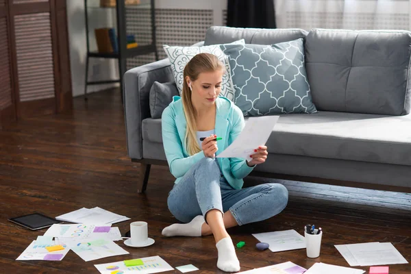 Junge Frau arbeitet mit Dokumenten, während sie zu Hause auf dem Fußboden sitzt. Studentin, Unternehmerin oder Freiberuflerin, die aus der Ferne über das Internet arbeitet oder studiert. — Stockfoto