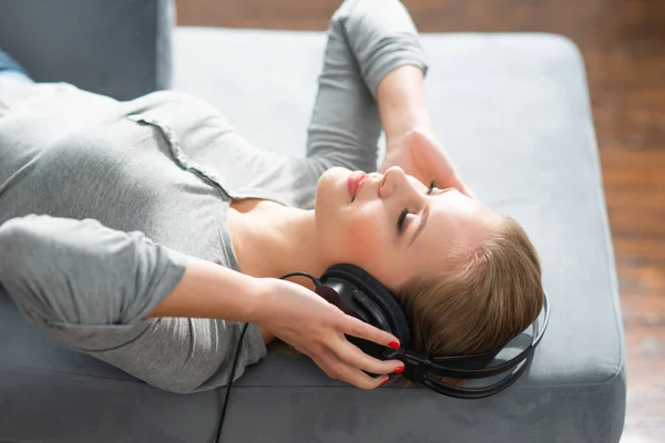 Ung vacker flicka ligger på en soffa i hörlurar och lyssnar på musik. Glad blond kvinna vilar hemma. — Stockfoto