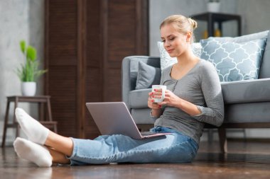 Genç bir kadın evde yerde otururken dizüstü bilgisayar kullanıyor. Öğrenci, girişimci veya serbest çalışan kız İnternet üzerinden çalışıyor veya uzaktan öğrenim görüyor.