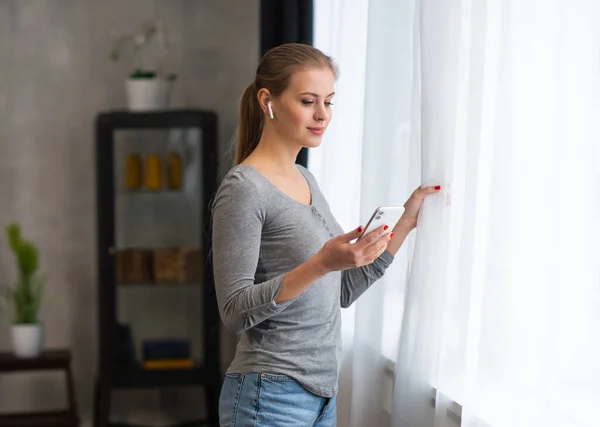Joven adolescente está de pie con un teléfono inteligente en frente de la ventana y mirando en él. Mujer rubia en casa. — Foto de Stock