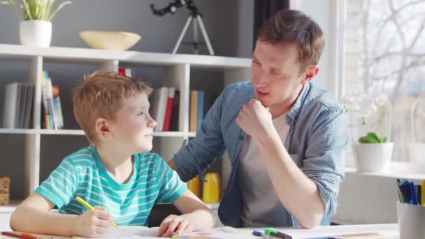 Το αγόρι και ο πατέρας κάνουν τις εργασίες στο τραπέζι. Το χαριτωμένο παιδί μαθαίνει στο σπίτι με τη βοήθεια των εγχειριδίων και των σχολικών υλικών. Η έννοια των σπουδών και της εκπαίδευσης. — Αρχείο Βίντεο