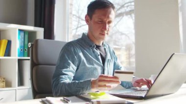 Genç adam internetten kredi kartıyla alışveriş yapıyor. Evde, bilgisayarın başında çalışan kişi. Çevrimiçi alışveriş ve ödeme sistemleri kavramı.