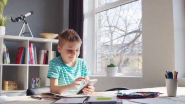 Çocuk Masa 'da ödevini yapıyor. Şirin Çocuk, Akıllı Telefon Cihazı ve Okul Eğitim Kitapları 'nın yardımıyla Evde Öğreniyor. Çalışma ve Eğlence Konsepti .