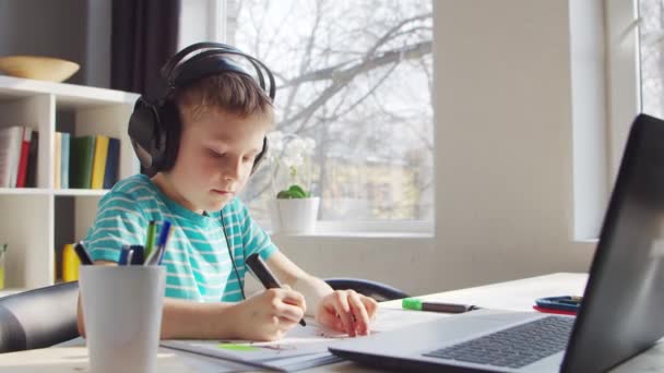 Băiatul îşi face temele la masă. Copilul drăguț învață acasă cu ajutorul laptopului și a manualelor școlare. Conceptul de studiu și educație la distanță. — Videoclip de stoc