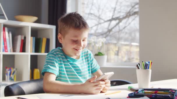 少年はテーブルで宿題をしている。かわいい子供はスマートフォンデバイスと学校教育教科書の助けを借りて自宅で学んでいます。勉強と娯楽の概念 . — ストック動画