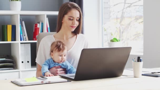 Junge Frau arbeitet, während sie zu Hause mit ihrem neugeborenen Baby sitzt. Arbeitsplatz der Unternehmerin, Freiberuflerin oder Studentin. Fernarbeit und Bildungskonzept. — Stockvideo