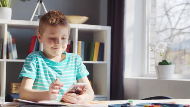 Το αγόρι κάνει τις εργασίες του στο τραπέζι. Χαριτωμένο παιδί μαθαίνει στο σπίτι με τη βοήθεια της συσκευής Smartphone και Σχολικής Εκπαίδευσης Εγχειρίδια. Μελέτη και ψυχαγωγία Concept . — Αρχείο Βίντεο