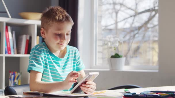 Boy fait ses devoirs à la table. Cute Child apprend à la maison à l'aide d'un téléphone intelligent et de manuels scolaires. Concept d'étude et de divertissement . — Video