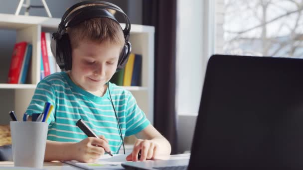 男孩正在餐桌前做作业。可爱的孩子在笔记本电脑和学校课本的帮助下在家学习。学习和远距离教育概念. — 图库视频影像
