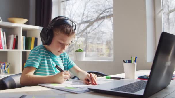 A fiú épp a házi feladatát végzi az asztalnál. Aranyos gyermek tanul otthon segítségével Laptop Számítógép és iskolai tankönyvek. Tanulmányi és távoktatási koncepció. — Stock videók
