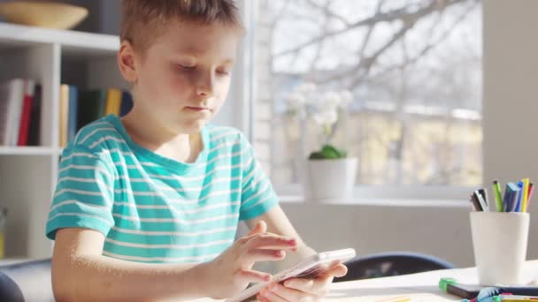 Boy fait ses devoirs à la table. Cute Child apprend à la maison à l'aide d'un téléphone intelligent et de manuels scolaires. Concept d'étude et de divertissement . — Video