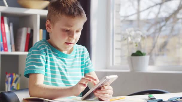 Хлопчик робить домашнє завдання за столом. Мила дитина вчиться вдома за допомогою смартфона пристрою та шкільних навчальних підручників. Концепція навчання та розваг  . — стокове відео