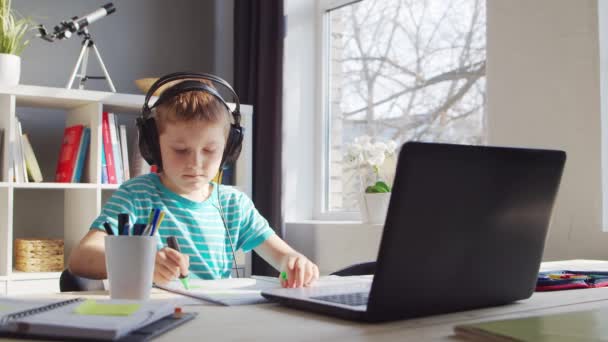Boy está haciendo su tarea en la mesa. Lindo niño está aprendiendo en casa con la ayuda de la computadora portátil y libros de texto de la escuela. Estudio y concepto de educación a distancia. — Vídeos de Stock