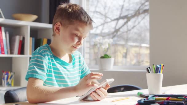 少年はテーブルで宿題をしている。かわいい子供はスマートフォンデバイスと学校教育教科書の助けを借りて自宅で学んでいます。勉強と娯楽の概念 . — ストック動画