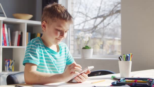 Pojken gör sina läxor vid bordet. Söta barn är att lära sig hemma med hjälp av Smartphone-enhet och skolans läroböcker utbildning. Studie- och underhållningskoncept . — Stockvideo