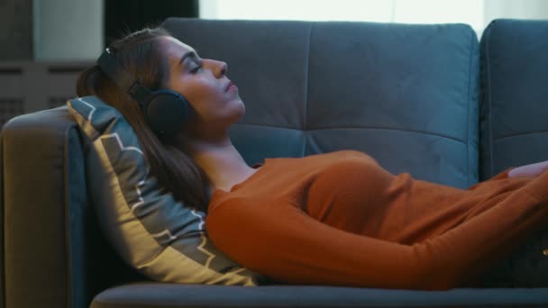 Młoda kobieta leży w domu na kanapie z zamkniętymi oczami i słucha muzyki na słuchawkach. Kobieta odpoczywa i słucha radia online. Relaks, medytacja i uważność. — Wideo stockowe
