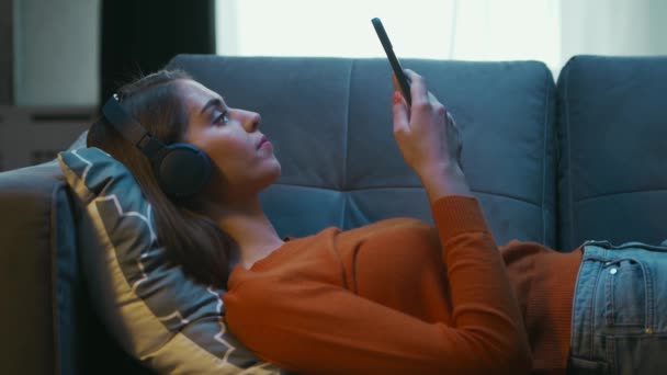 Молода жінка лежить вдома на каві зі своїм смартфоном Гаджет-пристроєм. Жінка відпочиває, розмовляє і прокручує газетні повідомлення. Розслаблення домівок і концепція соціальних медіа. — стокове відео