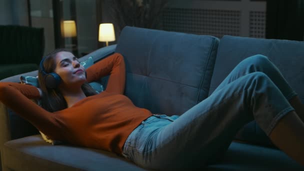 Молодая женщина лежит дома на диване с закрытыми глазами и слушает музыку в наушниках. Женщина отдыхает и слышит онлайн-радио. Отдых, медитация и памятование. — стоковое видео