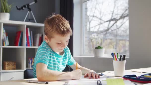 A fiú épp a házi feladatát végzi az asztalnál. Aranyos gyermek tankönyvek és iskolai anyagok segítségével otthon tanul. Tanulmányi és oktatási koncepció. — Stock videók