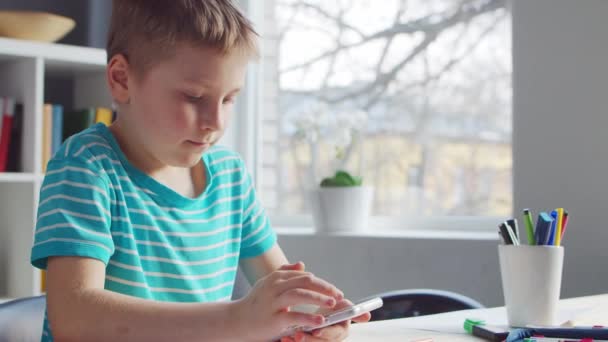 Boy fait ses devoirs à la table. Cute Child apprend à la maison à l'aide d'un téléphone intelligent et de manuels scolaires. Concept d'étude et de divertissement . — Video