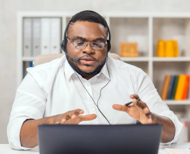 Afro-Amerikalı adam merkez ofiste bilgisayar, kulaklık ve diğer cihazları kullanarak çalışıyor. Eleman konferans görüşmesi yapıyor. Uzak görev.