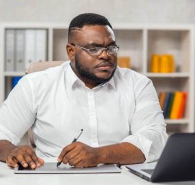 Ofisteki işyerinde. Genç Afro-Amerikalı bir adam bilgisayar, grafik tableti ve diğer cihazları kullanıyor. Uzak görev konsepti.