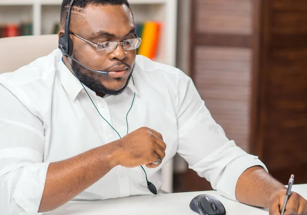 African-American man works at home office using computer, headset and other devices. Employee is having a conference call. Remote job. — Stock Photo, Image