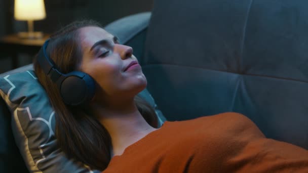 Young Woman está deitada em casa no sofá com seus olhos fechados e ouvindo música nos fones de ouvido. Mulher está descansando e ouvindo a Rádio Online. Relaxamento, Meditação e Mindfulness. — Vídeo de Stock