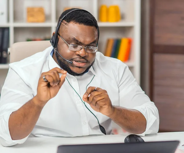 African-American man works at home office using computer, headset and other devices. Employee is having a conference call. Remote job. — ストック写真