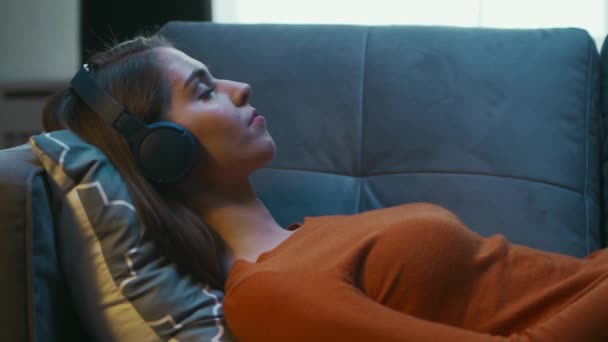 Young Woman está acostada en casa en el sofá con los ojos cerrados y escuchando música en los auriculares. La mujer está descansando y escuchando a la radio en línea. Relajación, Meditación y Mindfulness. — Vídeo de stock