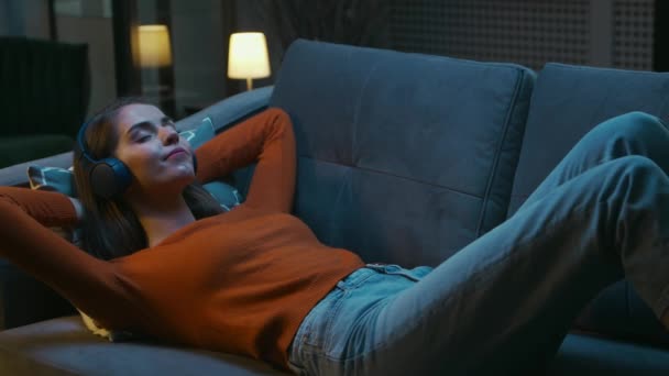Mladá žena leží doma na gauči se zavřenýma očima a poslouchá hudbu na sluchátkách. Žena odpočívá a poslouchá online rádio. Relaxace, meditace a bdělost. — Stock video