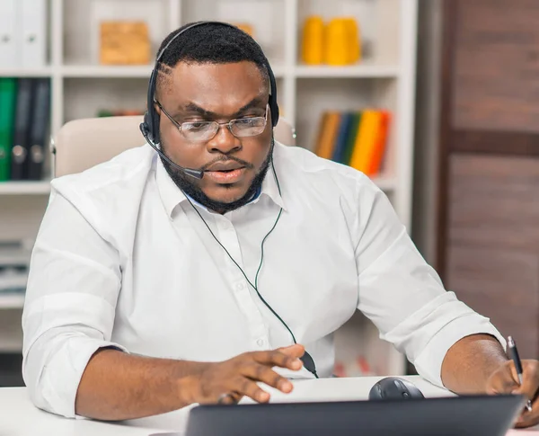 African-American man works at home office using computer, headset and other devices. Employee is having a conference call. Remote job. — ストック写真
