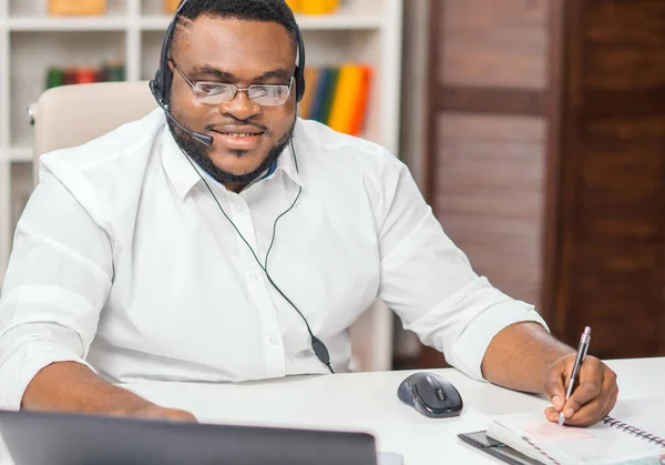 African-American man works at home office using computer, headset and other devices. Employee is having a conference call. Remote job. — ストック写真