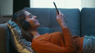 Genç Kadın Akıllı Telefon Gadget Cihazıyla Evdeki Koltukta Uzanıyor. Kadın Dinleniyor, Sohbet ediyor ve Haber Kaydırıyor. Ev Rahatlığı ve Sosyal Medya Kavramı.