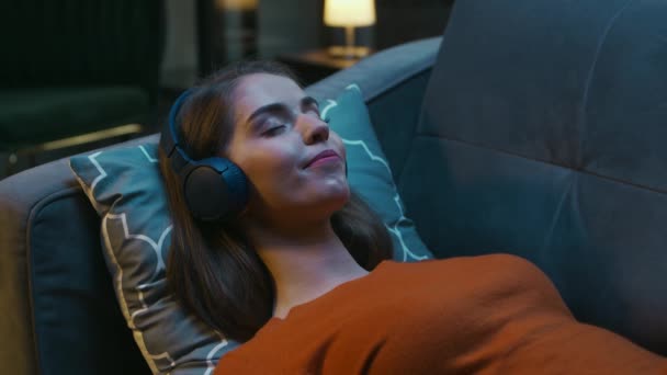 Unga kvinna ligger hemma på soffan med ögonen stängda och lyssnar på musik på hörlurar. Kvinnan vilar och hör till Online Radio. Avkoppling, meditation och mindfulness. — Stockvideo