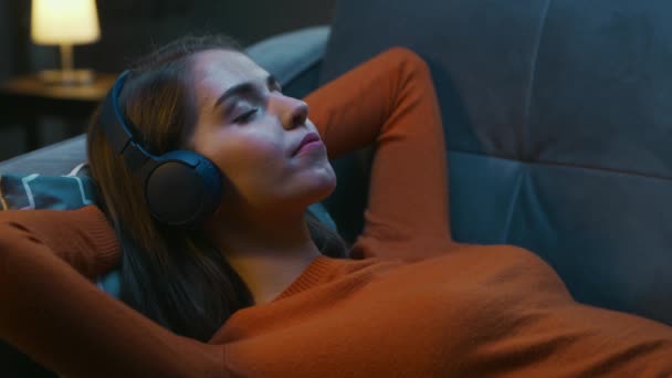 Die junge Frau liegt zu Hause mit geschlossenen Augen auf der Couch und hört Musik über Kopfhörer. Frau ruht sich aus und hört Online-Radio. Entspannung, Meditation und Achtsamkeit. — Stockvideo