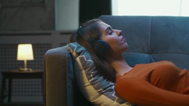 Молодая женщина лежит дома на диване с закрытыми глазами и слушает музыку в наушниках. Женщина отдыхает и слышит онлайн-радио. Отдых, медитация и памятование. — стоковое видео