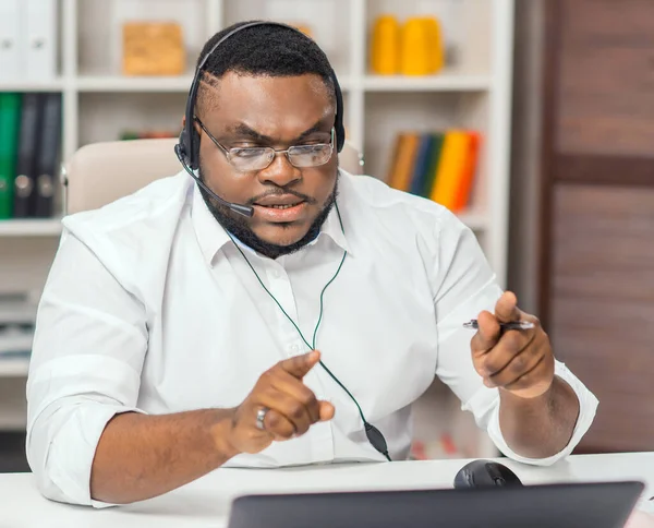 Afro-Amerikaanse man werkt op kantoor met behulp van computer, headset en andere apparaten. De werknemer heeft een conference call. Afstandsbediening. — Stockfoto