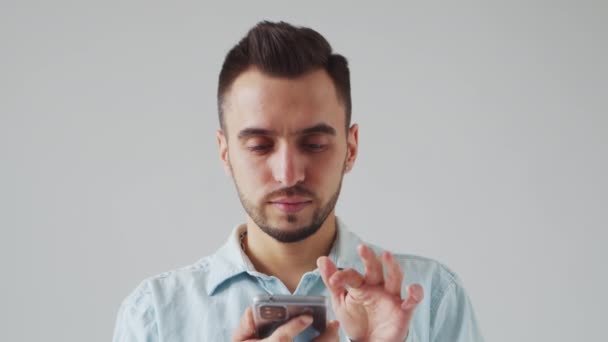 Young Caucasian Brunet Bearded Man with a Smartphone Device is Looking at the Camera on a Gray Background. Studio Male Face Portrait. Natural Day Light. — 비디오