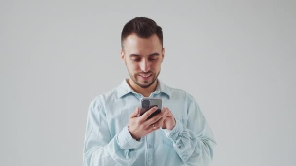 Giovane uomo bruno caucasico barbuto con un dispositivo Smartphone sta guardando la fotocamera su uno sfondo grigio. Studio Maschio Ritratto Volto. Luce naturale di giorno. — Video Stock