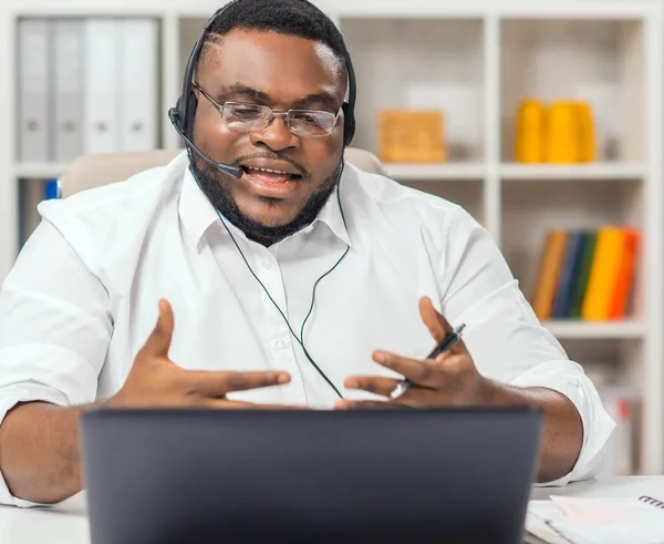 アフリカ系アメリカ人の男性は コンピュータ ヘッドセットなどのデバイスを使用して自宅のオフィスで働いています 従業員が電話会議を開いている リモートジョブコンセプト — ストック写真