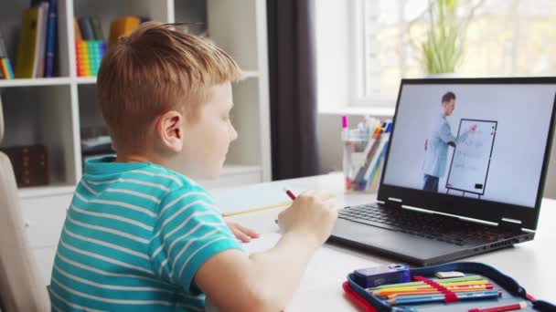Το αγόρι κάνει τις εργασίες του στο τραπέζι. Χαριτωμένο παιδί μαθαίνει στο σπίτι με τη βοήθεια του φορητού υπολογιστή και σχολικά εγχειρίδια. Μελέτη και εξ αποστάσεως Εκπαίδευση Έννοια. — Αρχείο Βίντεο