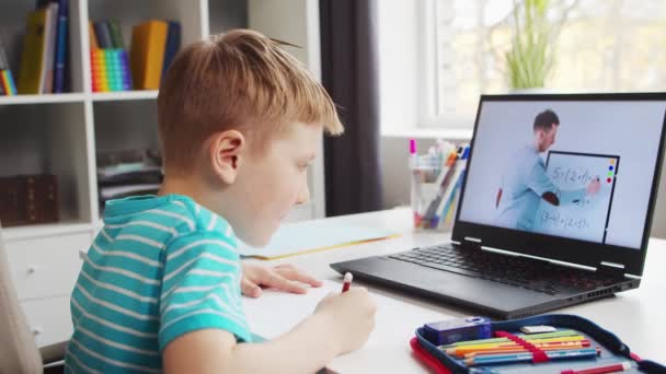 O rapaz está a fazer os trabalhos de casa na mesa. Criança bonito está aprendendo em casa com a ajuda de computadores portáteis e manuais escolares. Conceito de Estudo e Educação a Distância. — Vídeo de Stock