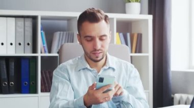 Young Caucasian Brunet Bearded Man with a Smartphone Device is Looking at the Camera on a Gray Background. Studio Male Face Portrait. Natural Day Light.