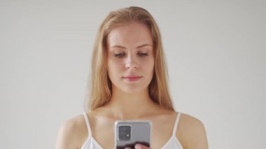 Genç, güzel, sarışın bir kadın kameranın önünde gri bir arka planda akıllı telefonuyla duruyor. Stüdyodaki çekici genç kızın portresi..