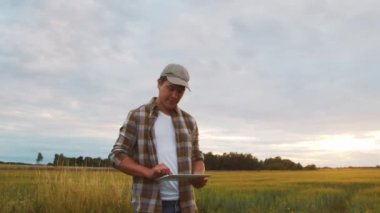 Farmer with a tablet computer in front of a sunset agricultural landscape. Man in a countryside field. The concept of country life, food production, farming and technology.