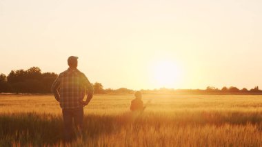Farmer and his son in front of a sunset agricultural landscape. Man and a boy in a countryside field. The concept of fatherhood, country life, farming and country lifestyle.