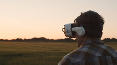 Farmer in virtual reality helmet in front of a sunset agricultural landscape. Man in a countryside field. The concept of country life, food production, farming and technology.
