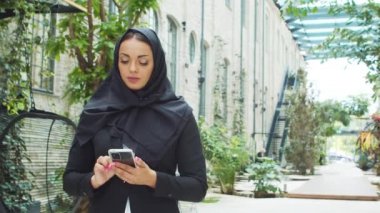 Portrait of young and confident Muslim business woman with a smartphone in hijab. Middle Eastern office worker or businesswoman is working outdoor in the city. Corporate and financial concept.